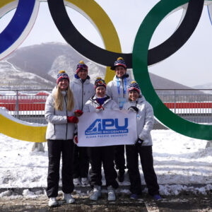 Skiers in front of Olympic circles with APU sign
